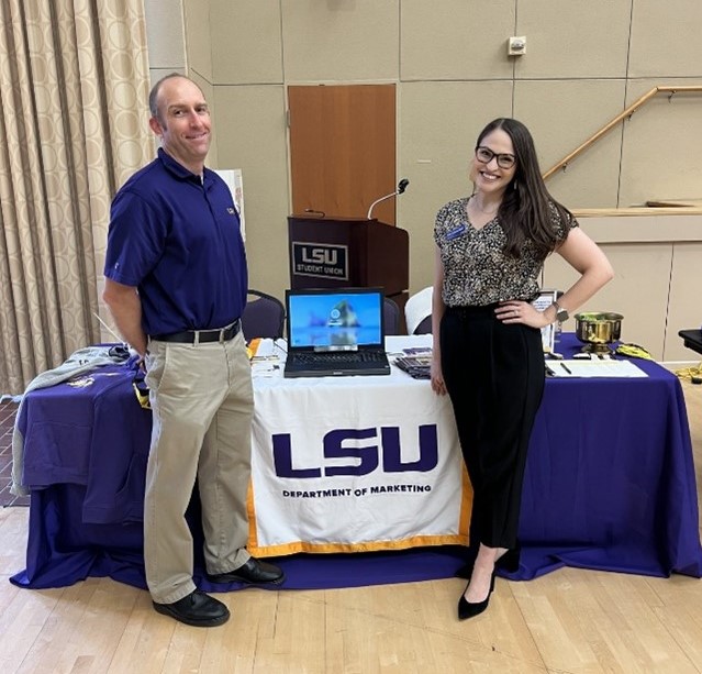 A male and female standing next to a table with a computer and other items on it. 