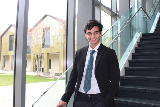 Samuel Camacho stands at the foot of a staircase in the BEC. He wears a suit and smiles at the camera.