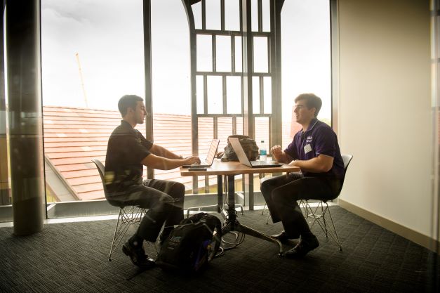 Two students talk at a table with laptops.